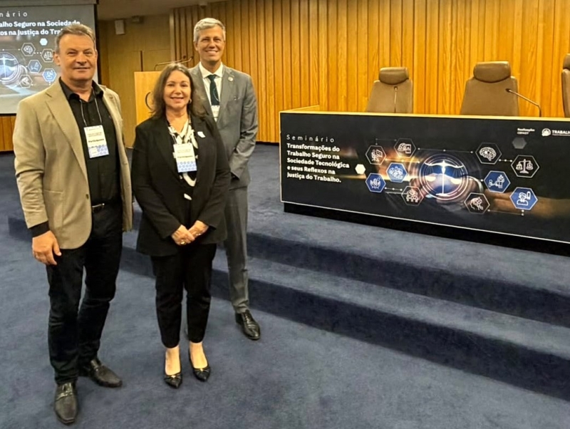 Dois homens e uma mulher posam para a foto. Ao lado deles, um banner com a identidade visual do evento