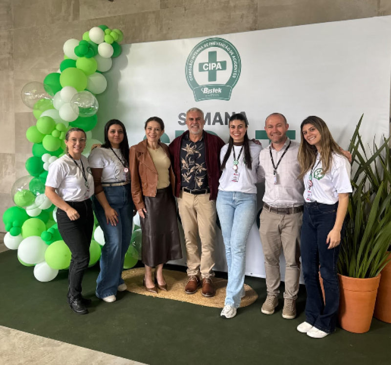 Cinco mulheres e dois homens em pé posam para foto, sobre um tapete verde escuro. Ao fundo, balões verde e branco decoram o ambiente