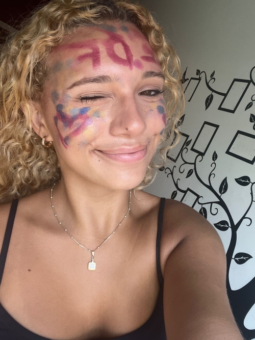 Aprendiz Beatriz, loira, cabelo crespo amarrado e com o rosto pintado, comemorando a aprovação no vestibular