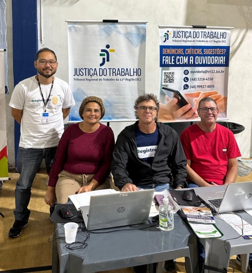 Três homens e uma mulher posam para foto em frente a duas mesas de plástico, com banners da Justiça do Trabalho atrás deles. Um dos homens está de pé.