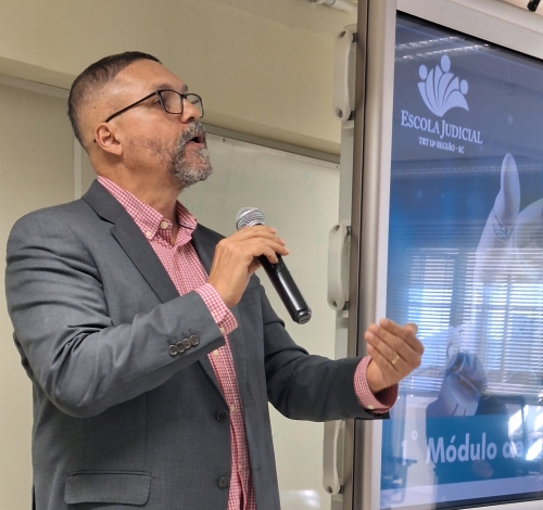 Homem de óculos, usando paletó cinza e camisa rosa, fala ao microfone e gesticula. Foto de perfil.