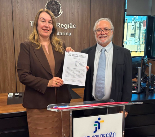 Uma mulher loira e um homem grisalho estão em pé, posando para a foto e exibindo um contrato assinado