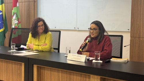 Duas mulheres em uma mesa de evento. Uma delas está com uma blusa vinho e fala ao microfone, sendo observada pela outra mulher, que veste uma blusa amarela