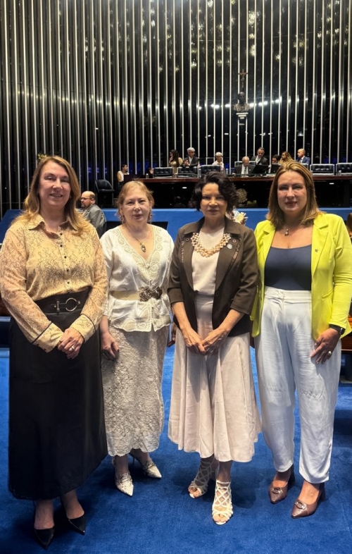 Quatro mulheres vestidas formalmente posam para foto, tendo ao fundo a mesa diretora do Senado Federal