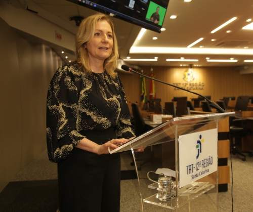 Mulher branca, loira, vestindo blusa preta com detalhes dourados, fala em um púlpito com a logomarca do TRT da 12ª Região