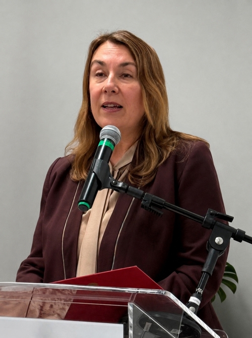 Busto de uma mulher branca, loira, vestindo blazer marrom e falando ao microfone