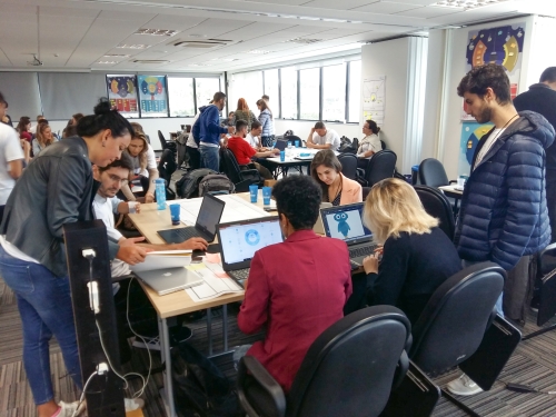 Equipe de seis pessoas trabalhando ao redor de uma mesa, com notebooks. Ao fundo, outras equipes também trabalham de forma semelhante.
