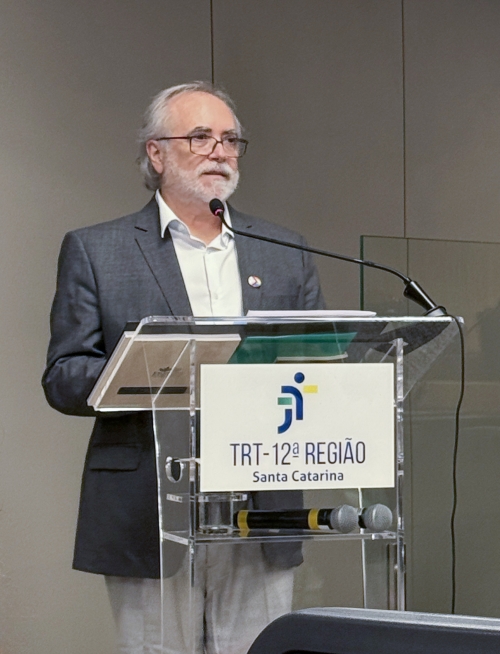 Homem branco de óculos, de cabelo e barba branca, fala ao microfone em um púlpito, onde aparece a logo do TRT da 12ª Região