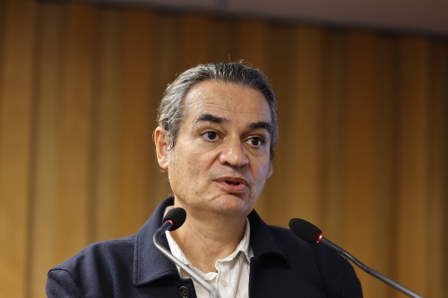 Homem de cabelo grisalho vestindo um terno com dois microfones na frente