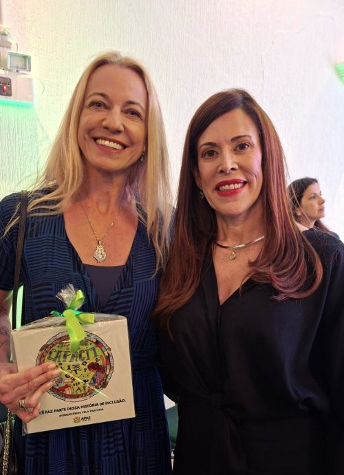 Uma mulher loira e outra ruiva posam para a foto, sorridentes. A loira exibe um pequeno presente com um laço verde
