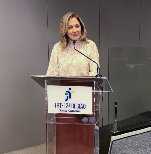 Mulher loira, cabelo até os ombros, vestindo blusa de renda de cor creme, fala ao microfone em um púlpito com a logomarca do TRT-SC