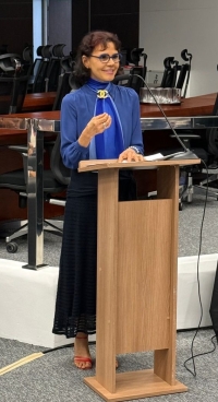 Mulher de óculos e vestido azul fala em um púlpito, ao microfone