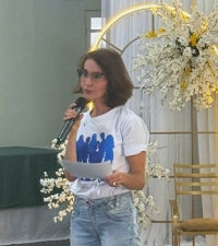 Mulher de óculos, com cabelos até os ombros, fala ao microfone segurando um papel