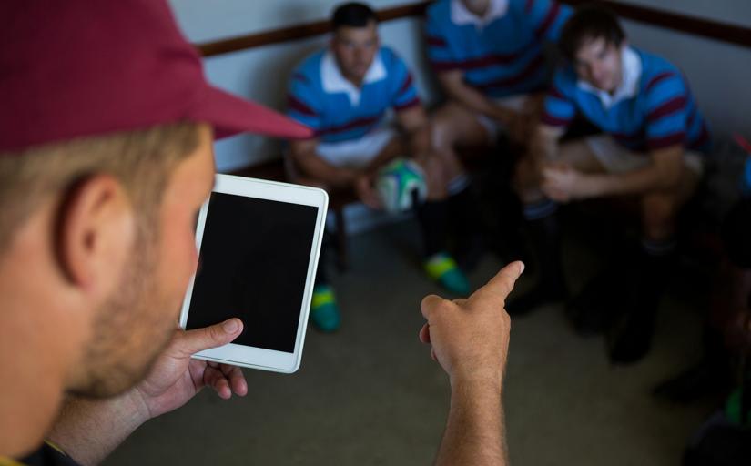 Fotografia de um técnico que segura um tablet e orientando jogadores em um vestiário