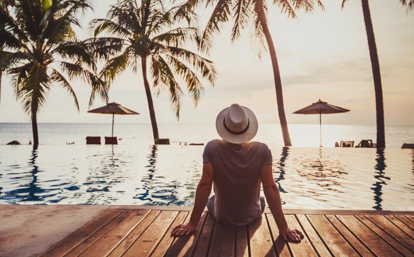 Homem sentado no deck, à beira do mar, de costas, olhando uma paisagem repleta de palmeiras