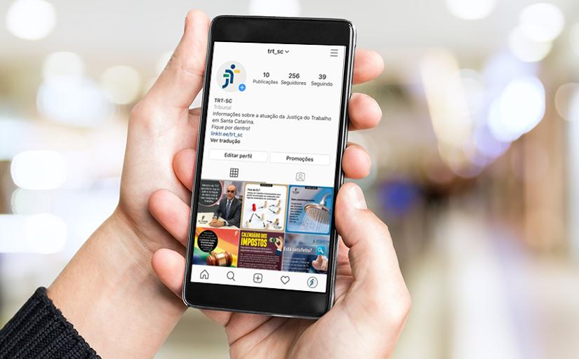 Foto de uma mão segurando um smartphone, que mostra em sua tela o perfil do TRT-SC no Instagram