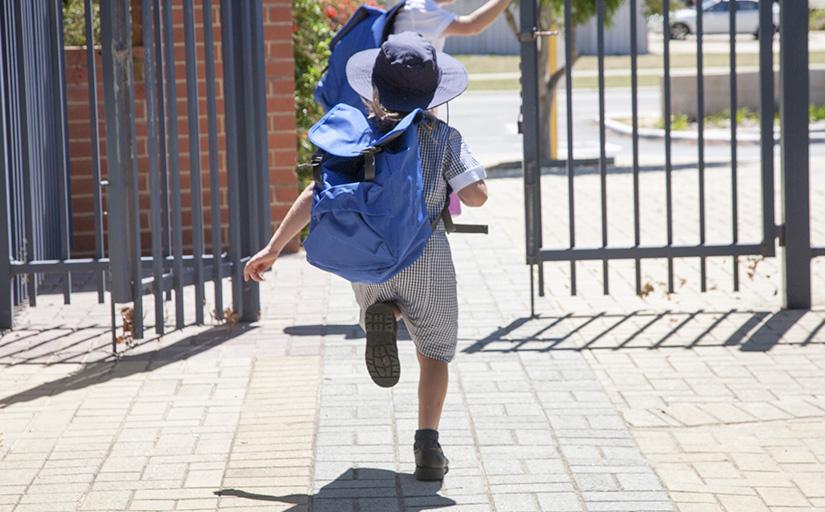 Foto mostra dois estudantes com mochilas correndo, saindo pelo portão de uma escola