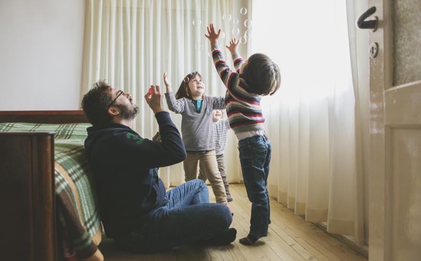 Pai brincando de bolhas de sabão com dois filhos no quarto
