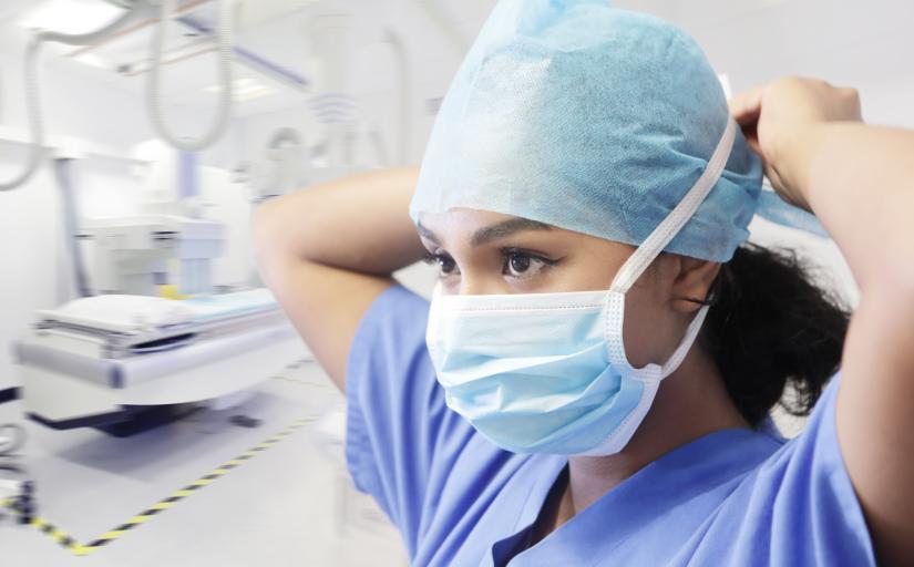 Profissional de saúde em primeiro plano, com touca azul, amarrando uma máscara cirúrgica de proteção