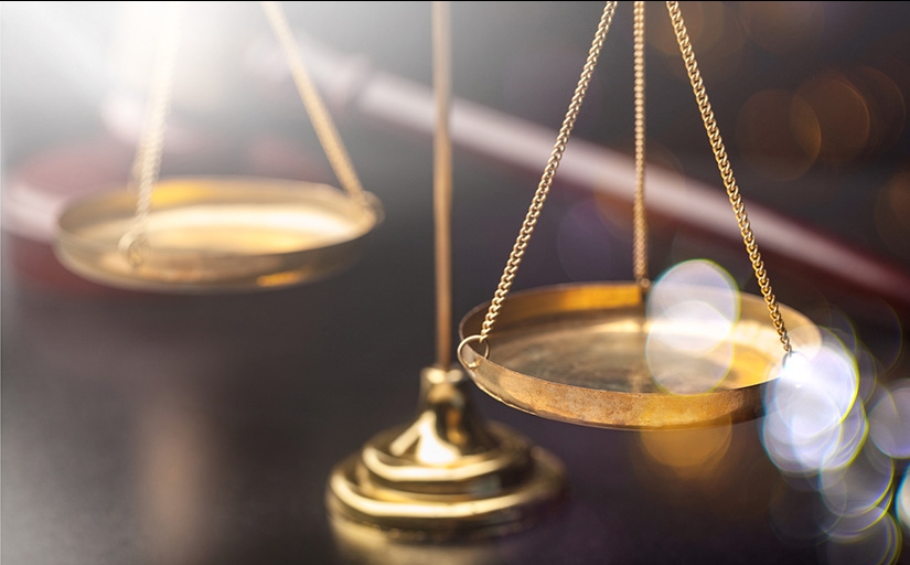Foto em close de uma balança dourada sobre uma mesa de madeira. A balança é um dos símbolos da Justiça e indica equilíbrio e ponderação na hora de pesar, lado a lado, os argumentos contra e a favor dos acusados.