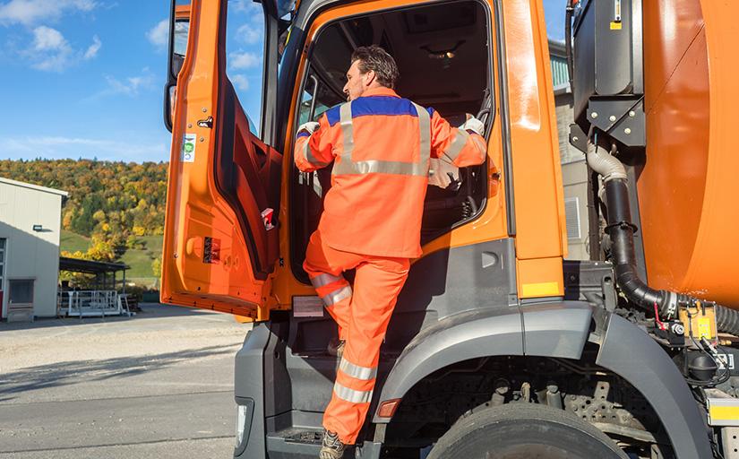 Foto ilustrativa mostra motorista com uniforme laranja embarcando em um caminhão de coleta de lixo urbano.