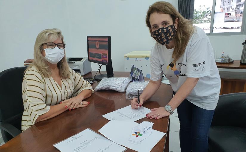 Secretária de Educação de Itajaí, sentada, à esquerda, juíza Andrea Waldrigues (de pé, à direita) posam para foto durante assinatura do termo de doação das máscaras. Ambas estão com máscaras de proteção.