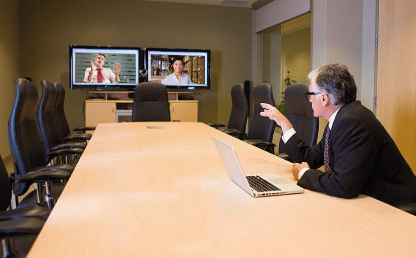 Homem sentado à mesa de reuniões falando com duas pessoas que aparecem em telas de TV, como numa videoconferência.