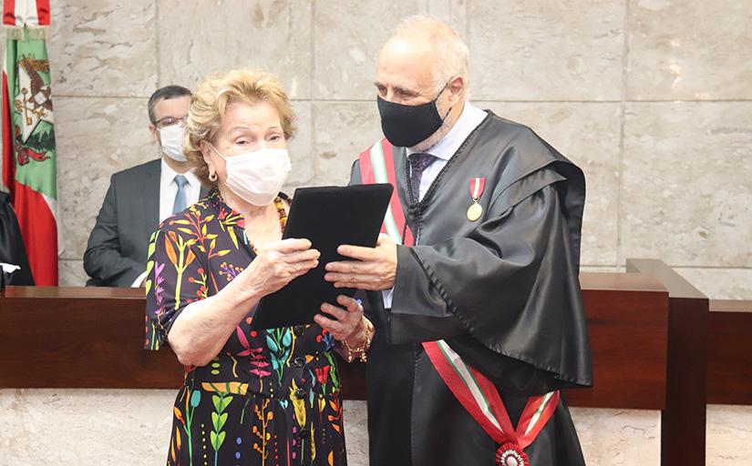Ione Ramos recebe placa das mãos de José Ernesto Manzi. Ambos estão de pé olhando para o objeto