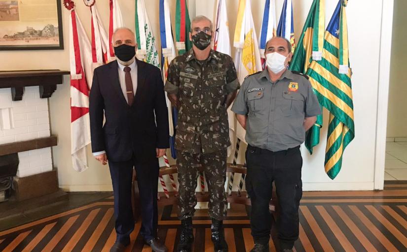 Três homens (um de terno, um de uniforme militar e outro de uniforme de segurança) posam para foto postados à frente de 12 bandeiras