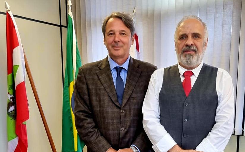 Um homem de terno e outro de colete e gravata posam sorridentes para a foto (cintura para cima), ao lado das bandeiras do Brasil e de Santa Catarina