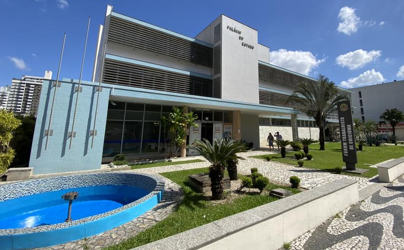 Fotografia da entrada do Fórum Trabalhista de Criciúma, um prédio de três andares com paredes pintadas nas cores branca e azul claro . É dia e o céu apresenta poucas nuvens