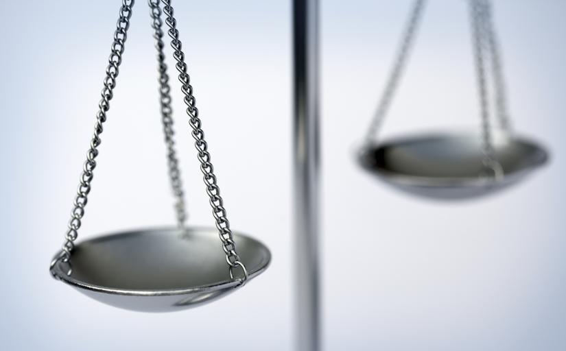 Fotografia ilustrativa de uma balança da justiça feita de metal. Ambas estão alinhadas, indicando igual distribuição de peso. 