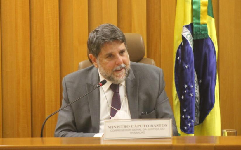 Ministro Caputo Bastos enquanto discursa sobre uma estrutura de madeira. Ao fundo, a brandeira do Brasil