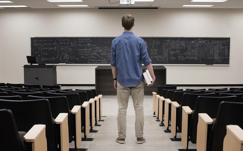 Homem jovem, de costas, em pé, observa uma lousa escura cheia de equações anotadas a giz, no corredor de uma sala de aula vazia