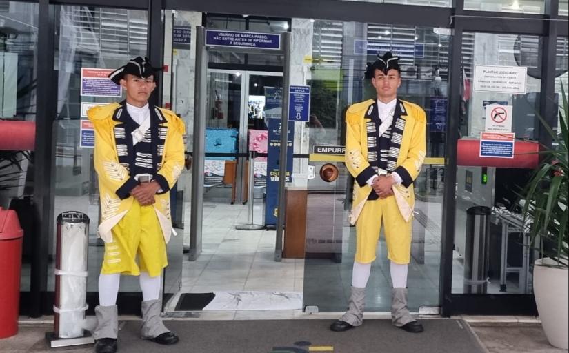 Dois soldados, em pé, na entrada do tribunal, vestem trajes históricos na cor amarela