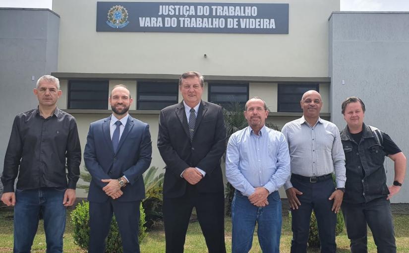 Seis homens vestindo trajes diversos posam para foto na frente de uma edificação que exibe a placa da Vara do Trabalho de Videira