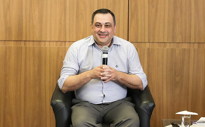Homem com camisa clara, sentado numa poltrona preta, segurando um microfone e sorrindo