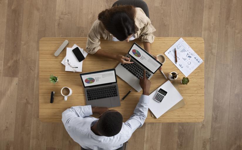 Foto de cima para baixo de uma mesa na qual uma mulher e um homem trabalhando em um computador