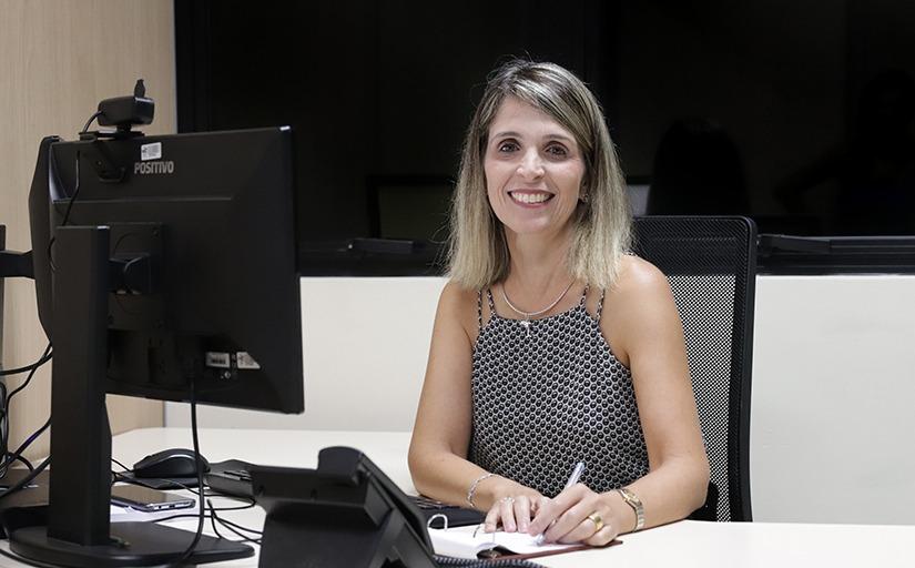 Mulher loira e sorridente, sentada na frente de um computador, com uma caneta em uma das mãos, escrevendo num bloco de papel