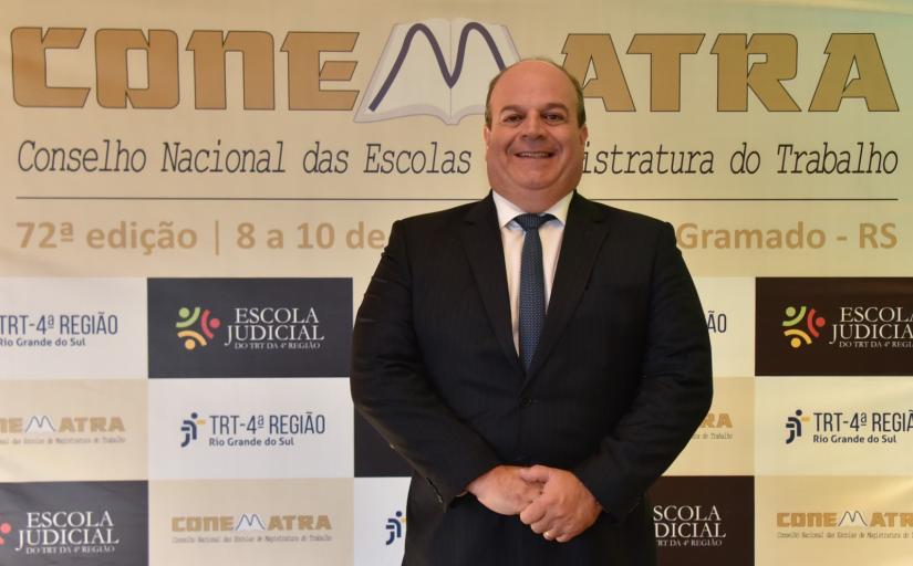 Homem branco, sorridente, de terno preto, posa para foto diante de um banner do evento Conematra