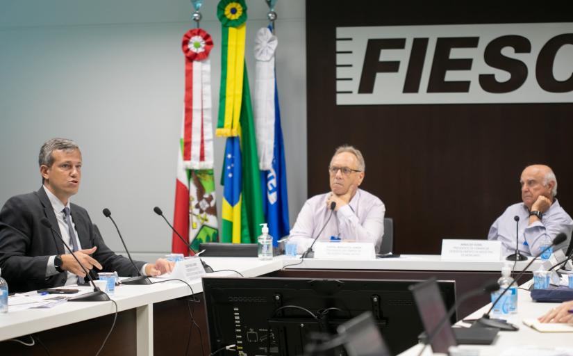 Três homens sentados atrás de uma mesa retangular, em formato de U. Ao fundo, bandeiras do Brasil uma logomarca escrito FIESC
