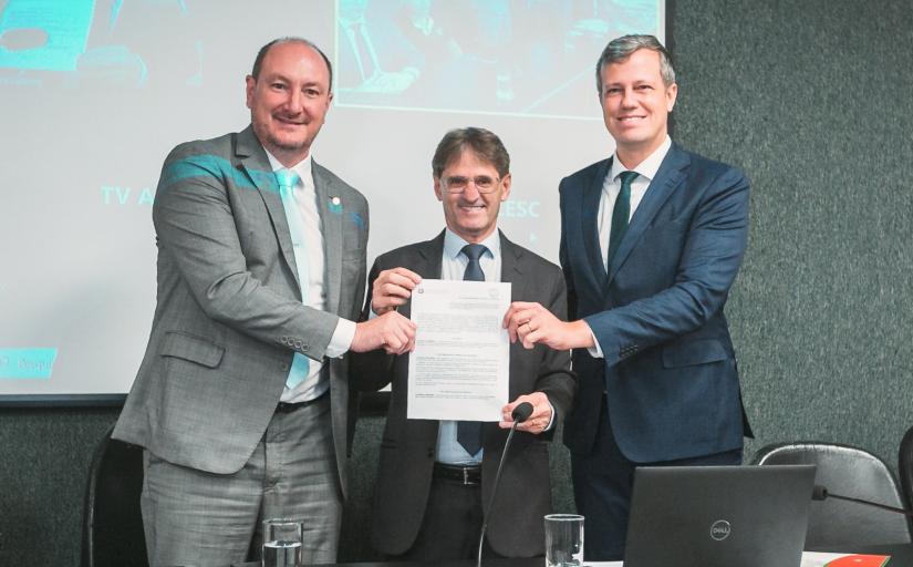 Foto com três homens de terno, em pé, sorridentes, segurando e mostrando um documento