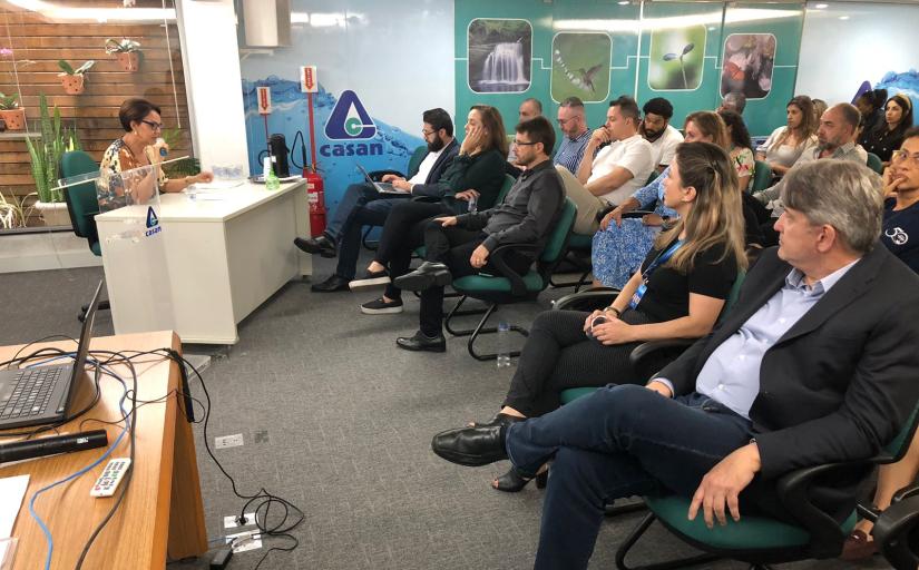 Foto com vista lateral de um auditório. Na imagem, auditório com cadeiras ocupadas e palestrante sentada à mesa falando ao microfone. 