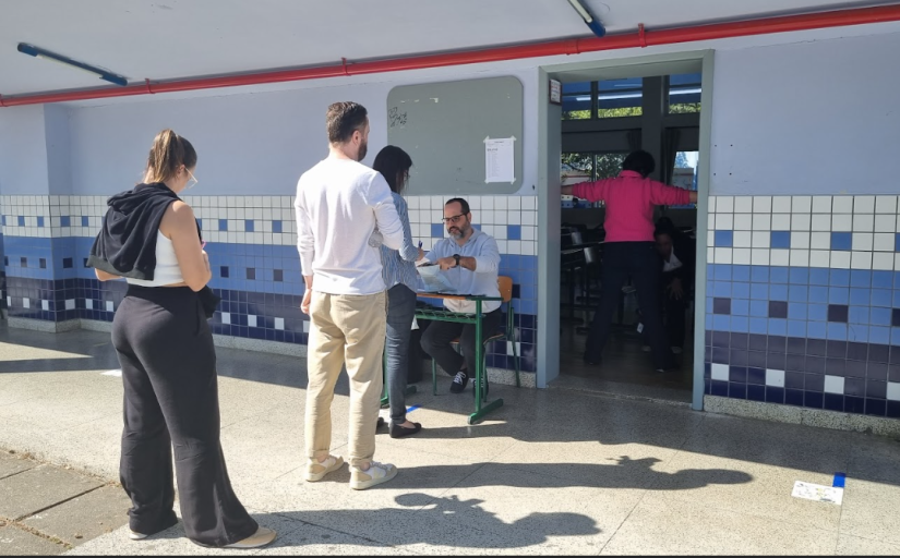 Foto mostra uma fila de pessoas aguardando para entrar em uma sala de aula. Uma delas está passando por um revista de detecção de metais