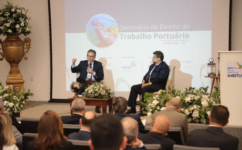 Foto tirada do fundo de um auditório com vista para o palco. Em primeiro plano está a plateia, em segundo plano, dois homens sentados no palco. Um deles fala ao microfone 