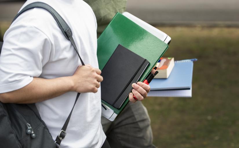 Fotografia ilustrativa em que aparece parte do tronco e das pernas de duas pessoas. Elas vestem roupas informais e caminham ao ar livre enquanto seguram mochilas e materiais escolares