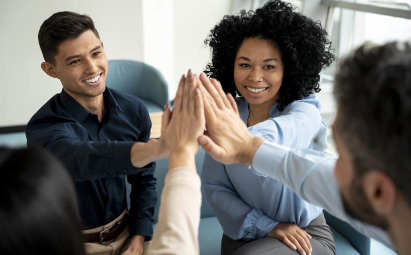 Foto mostra uma equipe de trabalho feliz aproximando as mãos no alto, comemorando um resultado