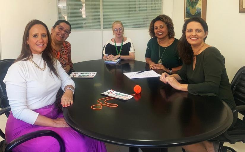 Grupo de cinco mulheres sentadas ao redor de uma mesa escura posam sorridentes para foto.