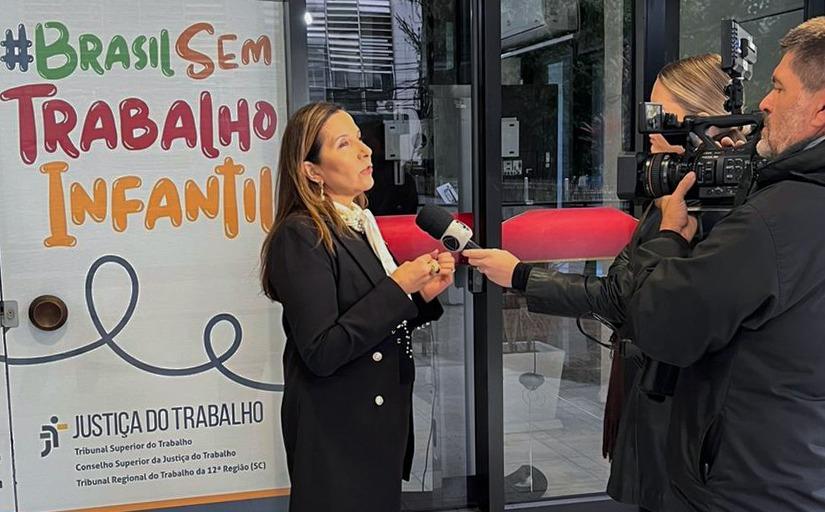 Fotografia de uma mulher sendo entrevistada para a televisão. Ela fala em um microfone enquanto uma câmera a filma. Atrás dela, há portas de vidro e em um é possível ler #Brasil Sem Trabalho Infantil