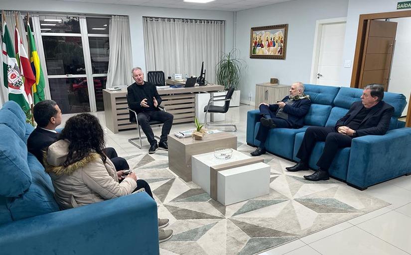 Quatro homens e uma mulher conversam sentados em sofás azuis. Um dos homens roupa preta está ao centro, falando